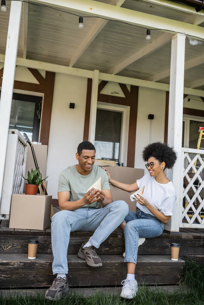 Pozitif Afro-Amerikalı çift, ellerinde sandviçler ve kahveyle yeni evin verandasında konuşuyorlar. - Fotoğraf, Görsel