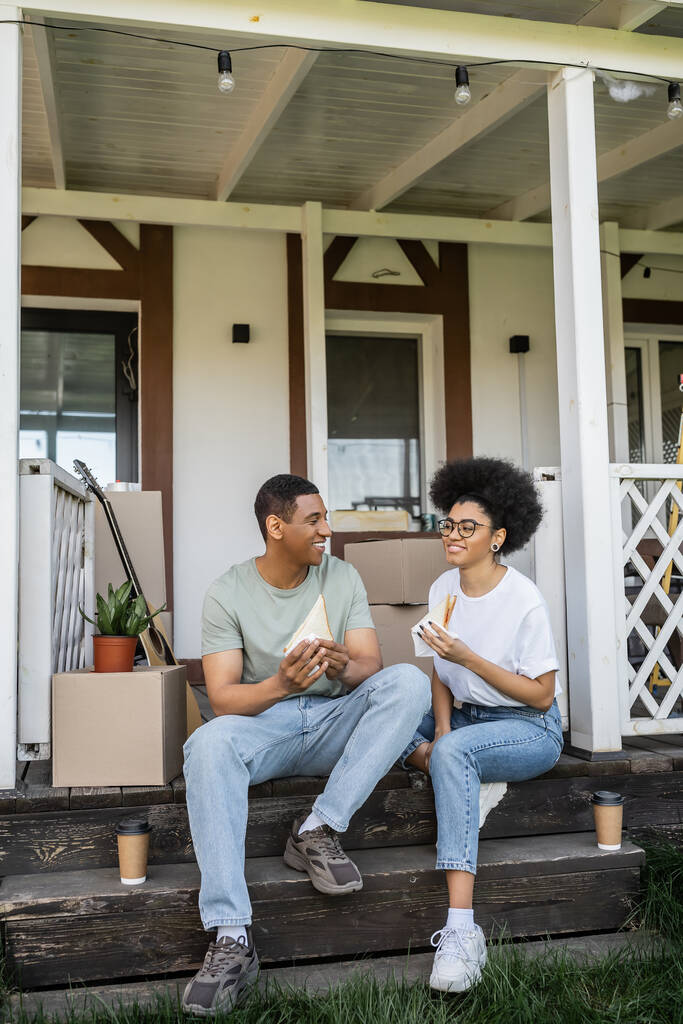 Mutlu Afro-Amerikalı kadın erkek arkadaşının yanında sandviç ve kahve tutuyor. Yeni evin verandasına gitmek için. - Fotoğraf, Görsel