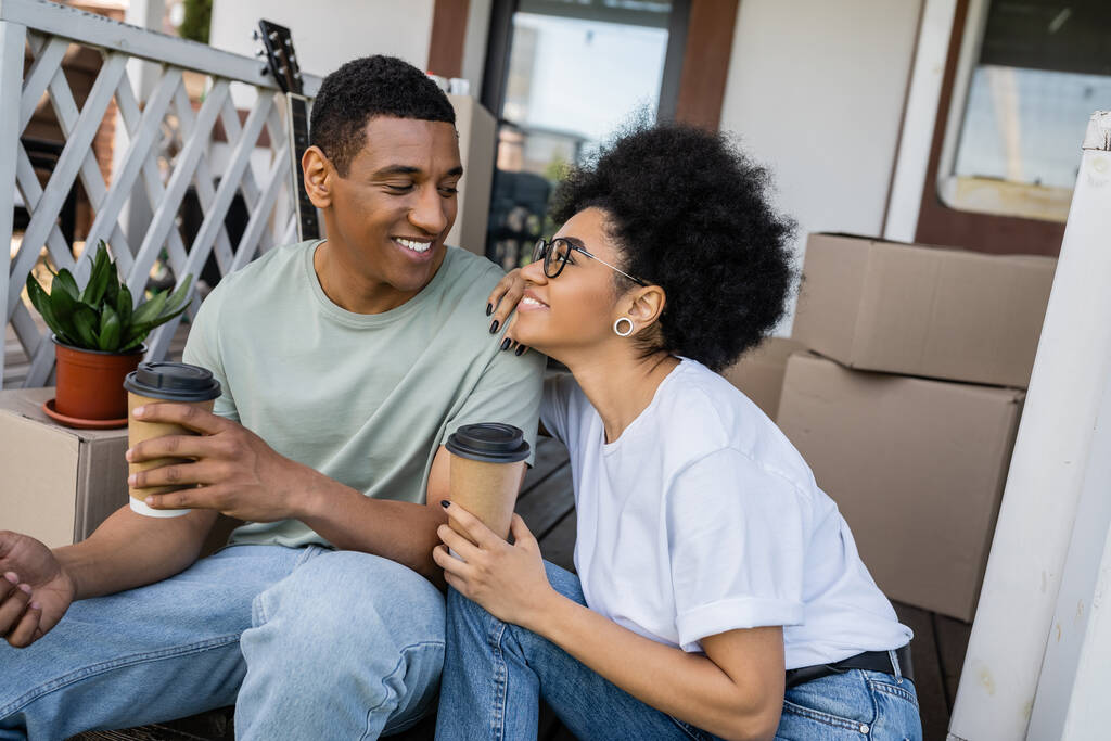 χαμογελαστό ζευγάρι Αφροαμερικανών κρατώντας καφέ και μιλώντας στη βεράντα του νέου σπιτιού - Φωτογραφία, εικόνα