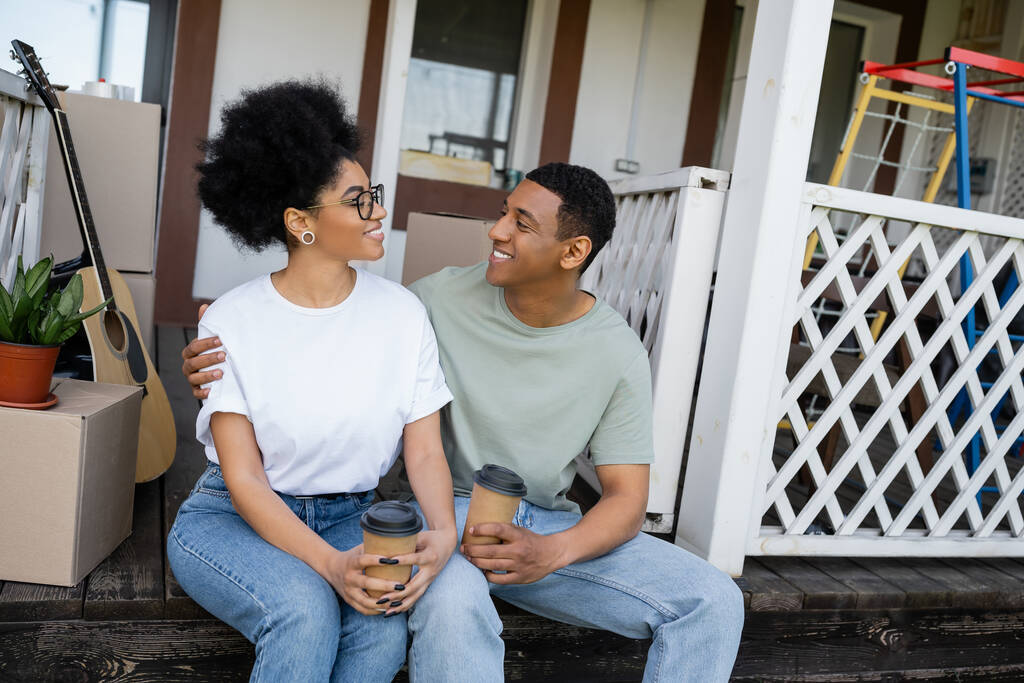 positief Afrikaans Amerikaans paar knuffelen en vasthouden van koffie te gaan in de buurt van dozen op veranda van nieuw huis - Foto, afbeelding