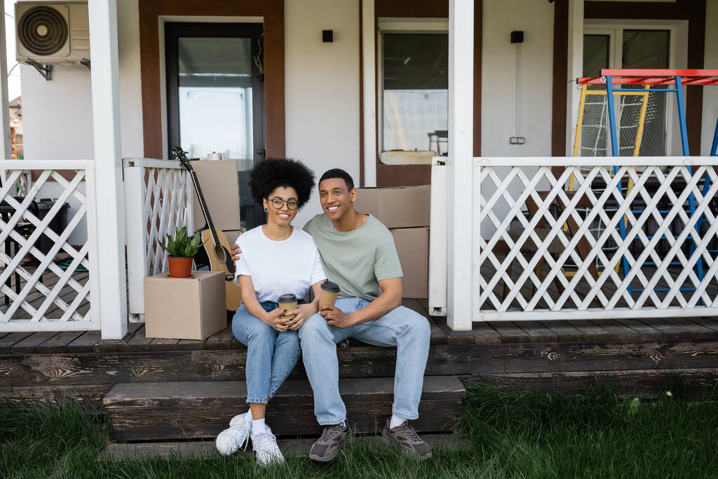 Gülümseyen Afrikalı Amerikalı çift ellerinde kahveyle yeni evin verandasındaki paketlerin yanına gidiyorlar. - Fotoğraf, Görsel