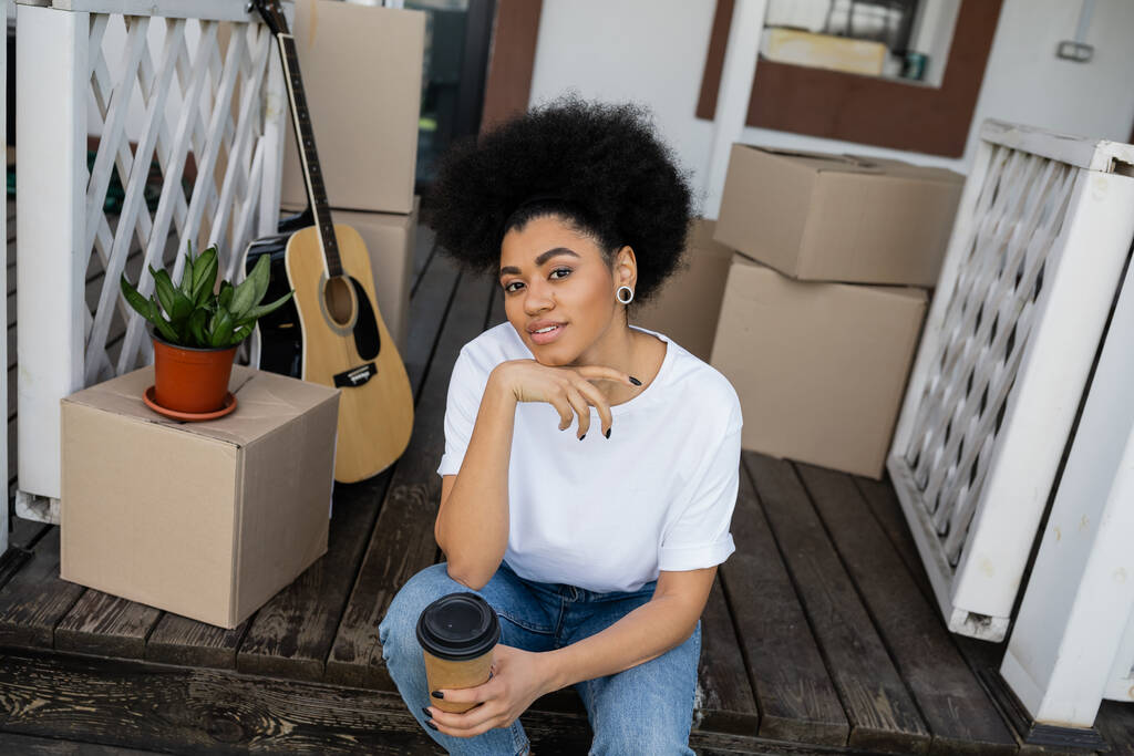 femme afro-américaine souriante tenant du café pour aller s'asseoir près des boîtes en carton et de la nouvelle maison - Photo, image