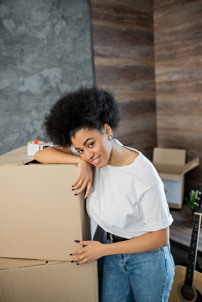 mulher americana africana alegre de pé perto de caixas de papelão e fita adesiva na sala de estar - Foto, Imagem