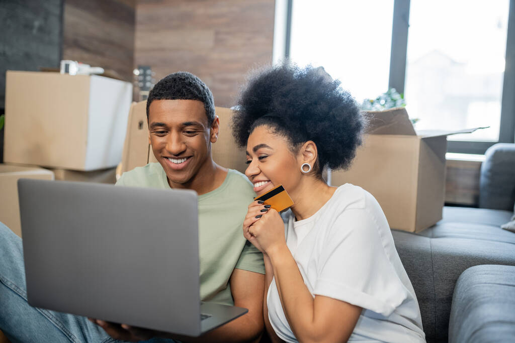 pareja afroamericana positiva utilizando el ordenador portátil y la tarjeta de crédito durante la reubicación en la nueva casa - Foto, Imagen
