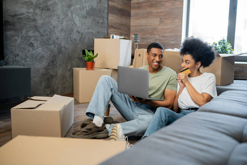 улыбающаяся африканская американская пара с помощью кредитной карты и ноутбука возле картонных коробок в новом доме - Фото, изображение