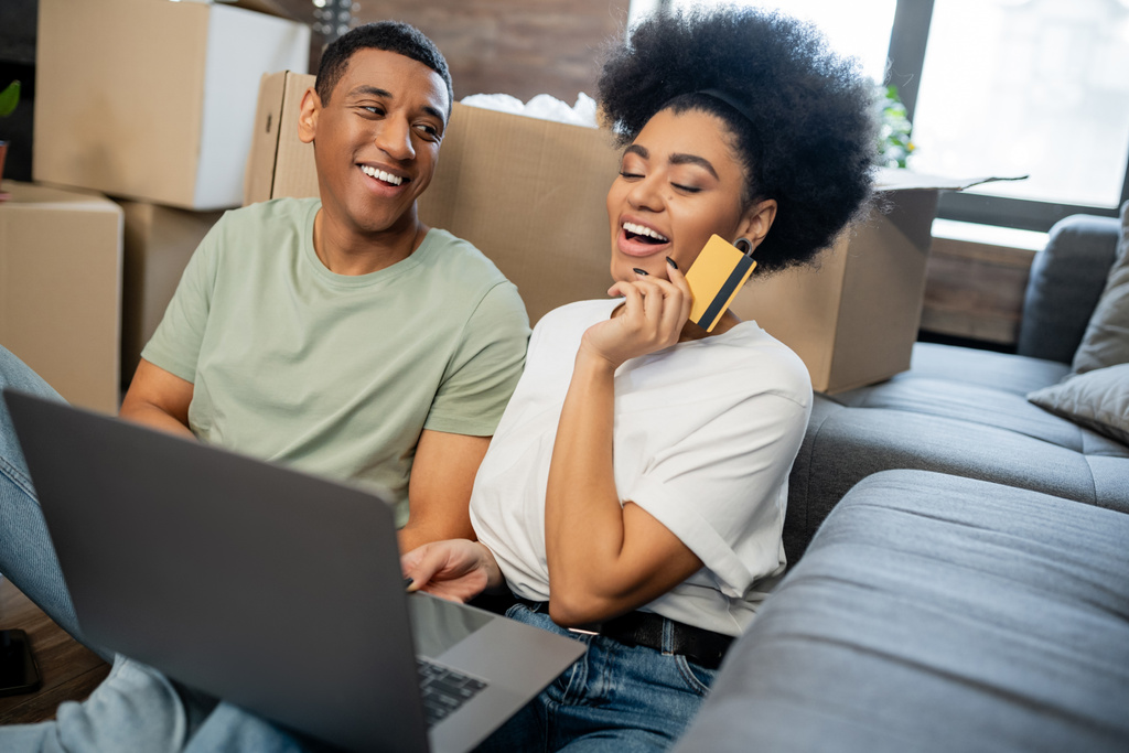 excitée femme afro-américaine faisant des achats en ligne près de petit ami et boîtes en carton dans une nouvelle maison - Photo, image