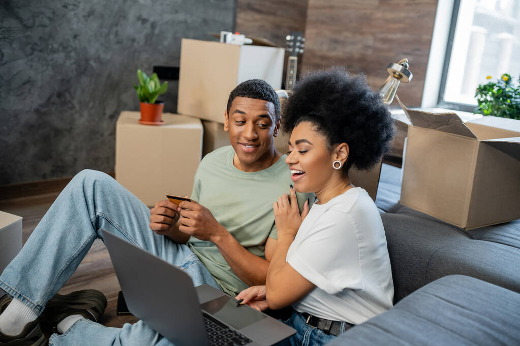carefree african american woman hugging boyfriend with laptop and credit card near boxes at home - Photo, Image