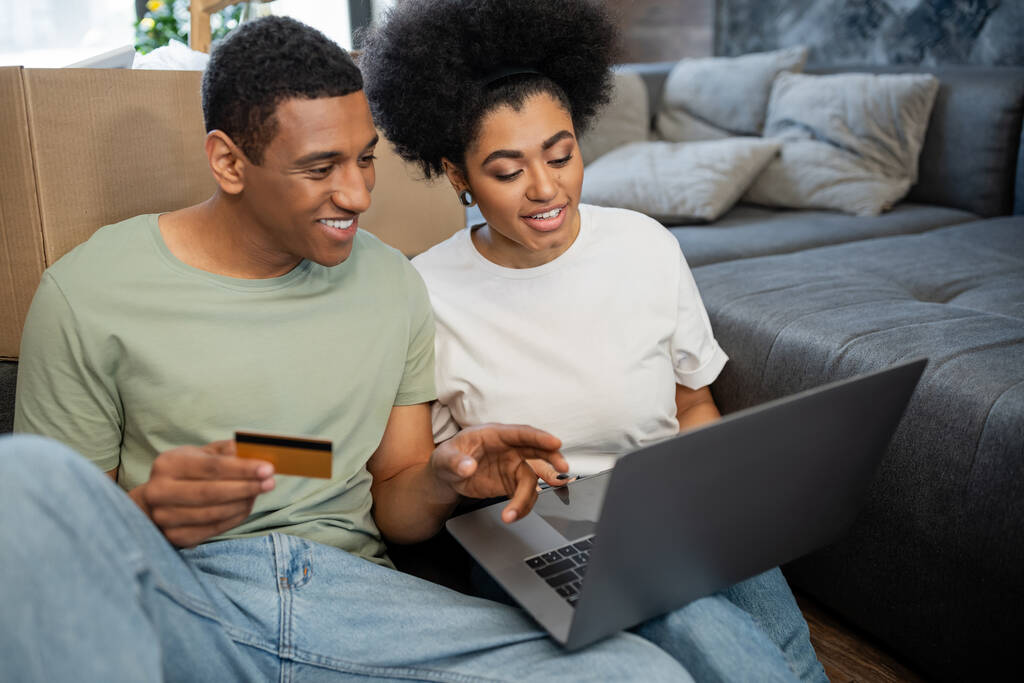 african american couple smiling during online shopping near carton box in living room in new house - Photo, Image