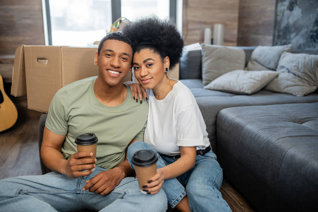 sonriente pareja afroamericana sosteniendo café para ir cerca de caja de cartón en nuevo salón - Foto, imagen