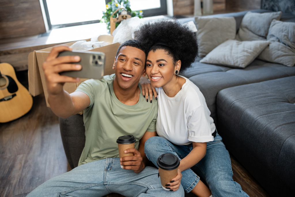 smiling african american couple with coffee taking selfie on smartphone near boxes in new house - Photo, Image