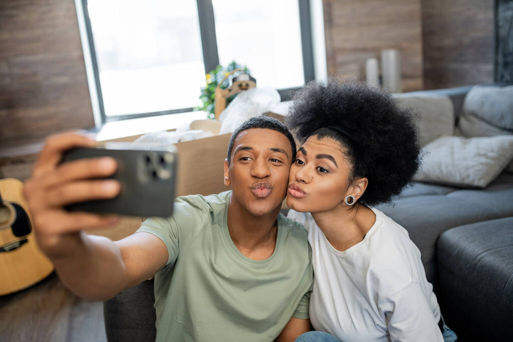pareja afroamericana haciendo pucheros labios mientras toma selfie en el teléfono inteligente en la nueva sala de estar - Foto, imagen