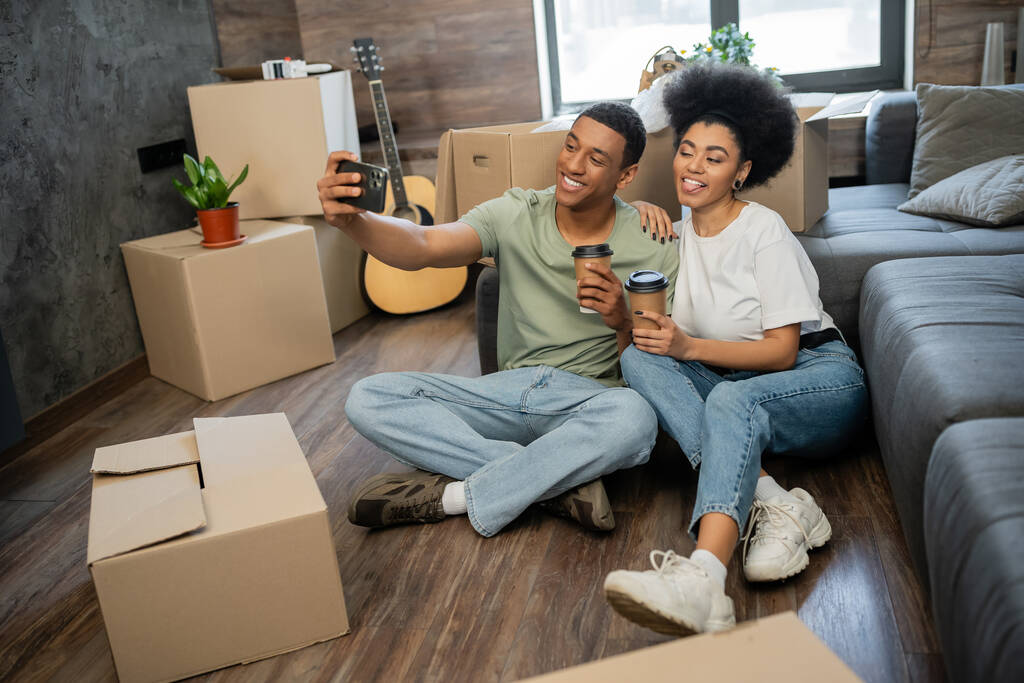 pareja afroamericana haciendo muecas y tomando selfie con café cerca de cajas en casa nueva - Foto, Imagen