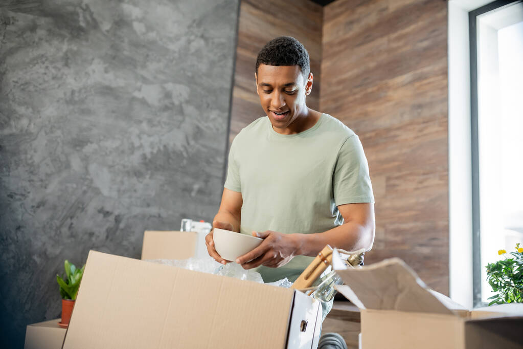joyful african american man holding plate while unpacking cardboard boxes in new house - Photo, Image