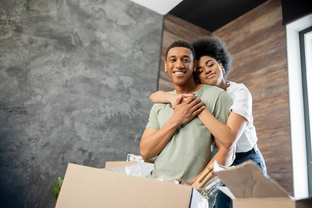 Gülümseyen Afrikalı Amerikalı kadın yeni evdeki paketlerin yanında dururken erkek arkadaşına sarılıyor. - Fotoğraf, Görsel