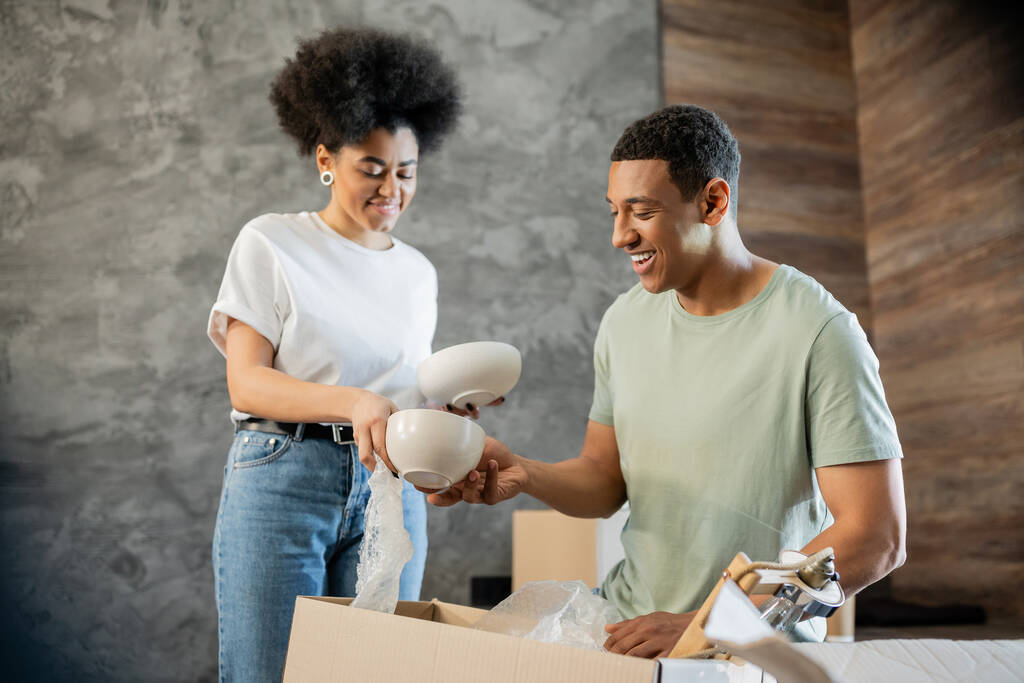 笑顔のアフリカ系アメリカ人の男がガールフレンドにテーブルウェアを与えながら,新しい家で箱を荷造りしている - 写真・画像
