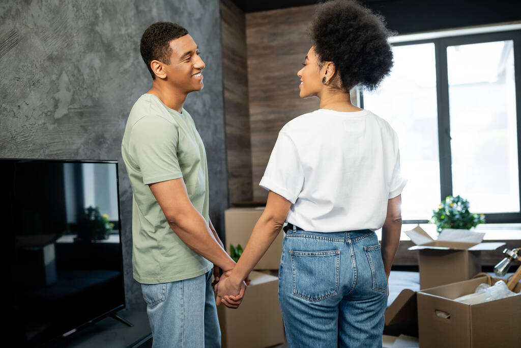 pareja afroamericana llena de alegría cogida de la mano mientras está de pie cerca de cajas de cartón en casa nueva - Foto, imagen