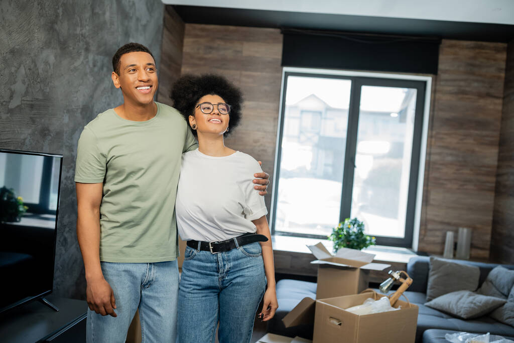 pareja afroamericana soñadora y sonriente abrazándose cerca de cajas de caron en casa nueva - Foto, Imagen
