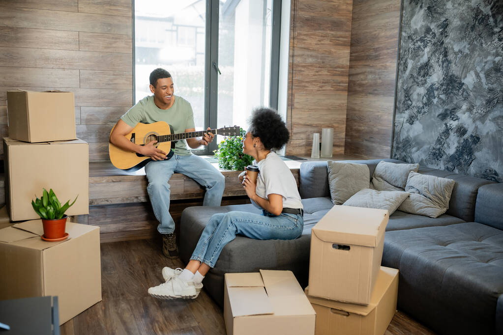 alegre pareja afroamericana con café tocando guitarra acústica cerca de cajas en casa nueva - Foto, Imagen