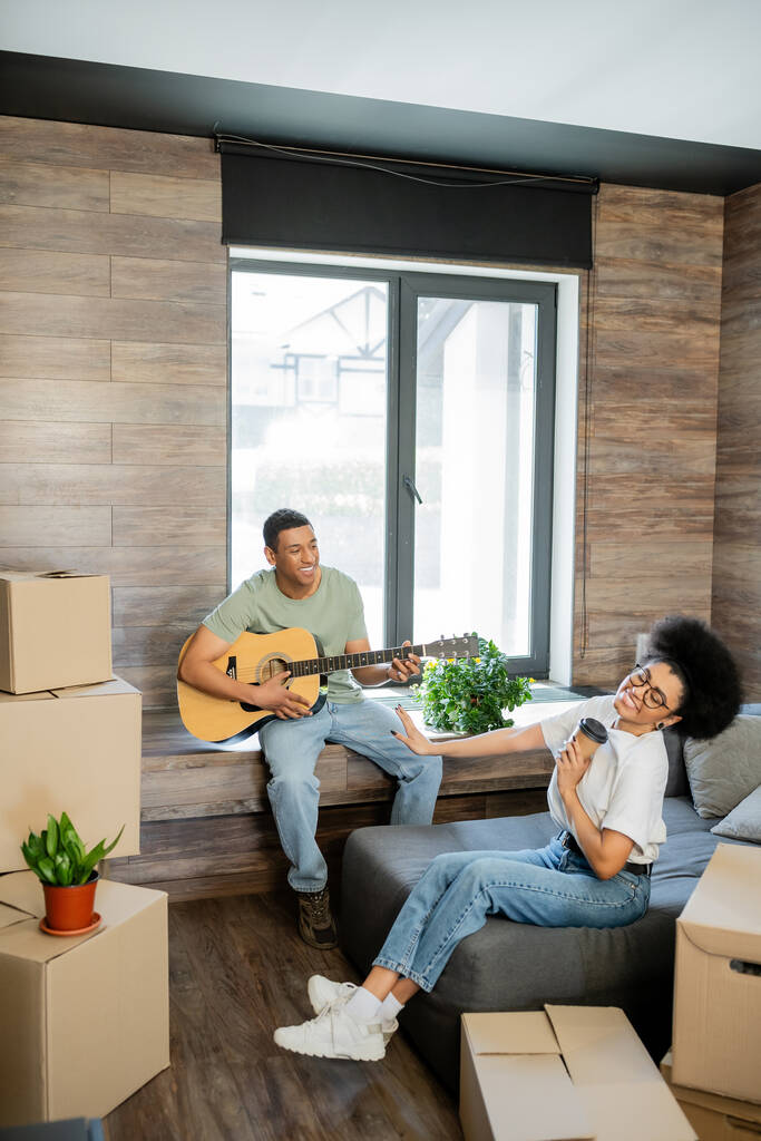 felice donna afroamericana in possesso di caffè vicino fidanzato suonare la chitarra acustica in una nuova casa - Foto, immagini