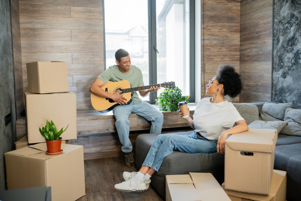 heureux homme afro-américain jouer de la guitare acoustique près de petite amie avec café à emporter dans une nouvelle maison - Photo, image