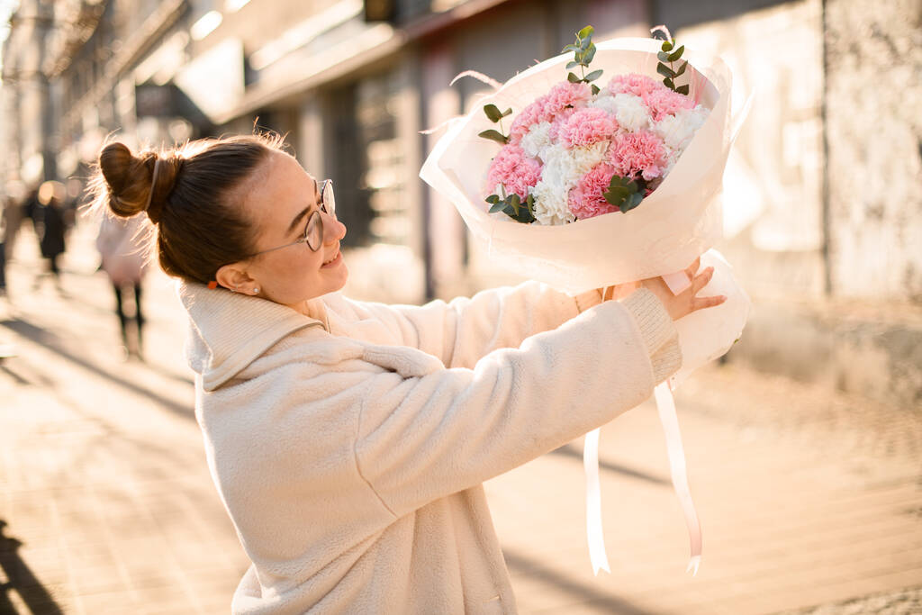 Çiçekçi genç bir kadın ortaya çıkıyor. Çiçek açan pembe, beyaz karanfiller ve okaliptüs dalları şehrin bulanık arka planında beyaz ambalaj kağıdında oluşturuluyor. - Fotoğraf, Görsel