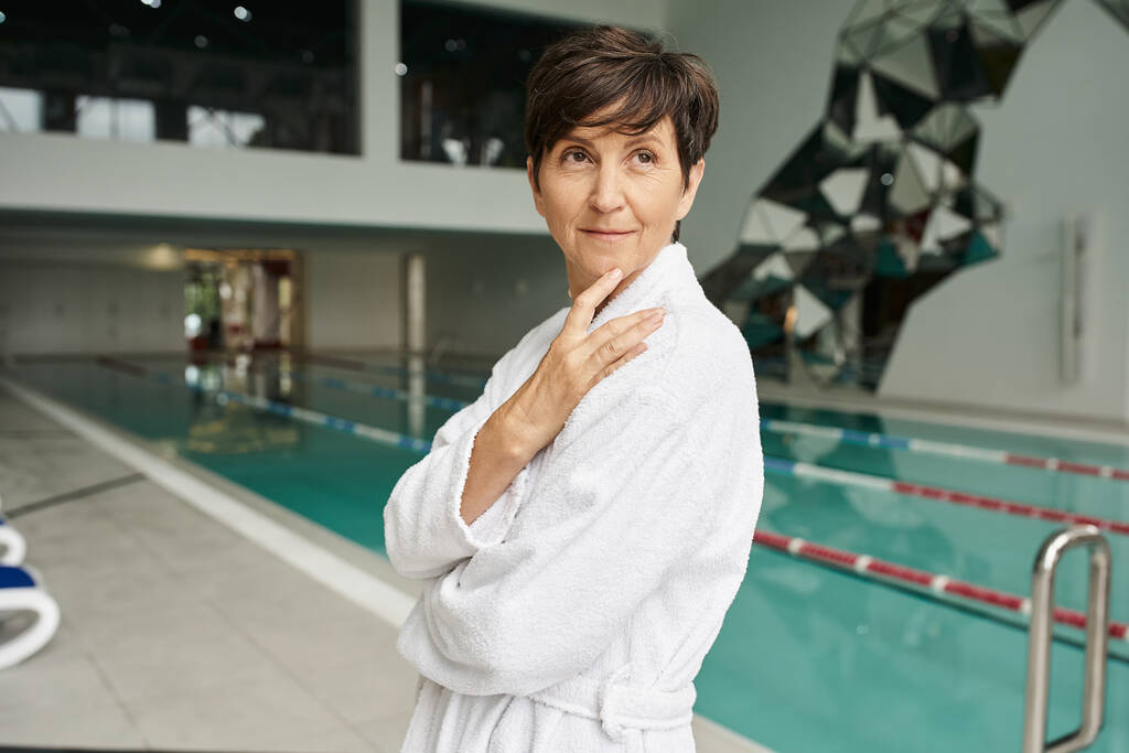 pleased middle aged woman with short hair standing in white robe, swimming pool, sport, smile - Photo, Image