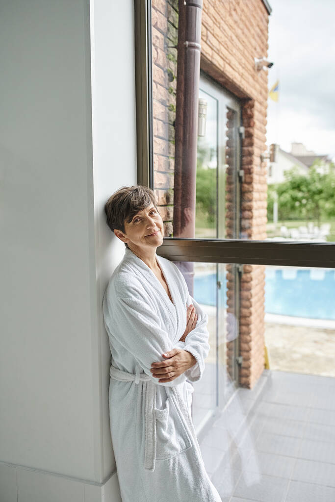 mujer madura de pie en bata cerca de la ventana panorámica en el centro de spa, vista a la piscina, mira a la cámara - Foto, imagen