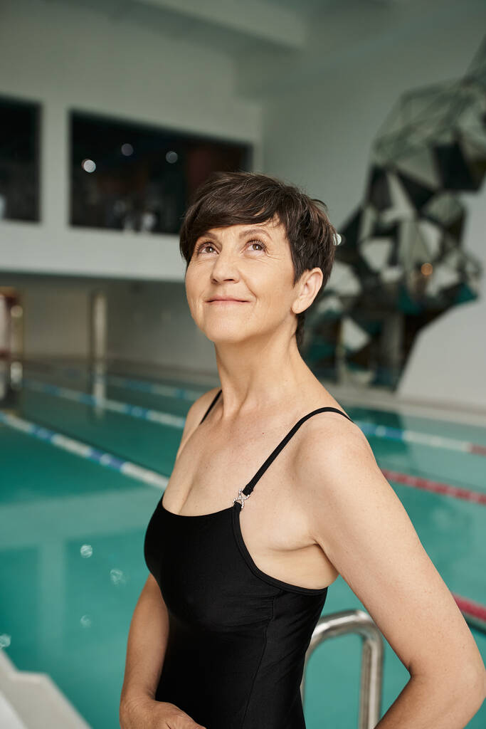 мечтательная женщина среднего возраста с короткими волосами позирует в купальнике рядом с крытым бассейном, спа-центр, смотреть вверх - Фото, изображение