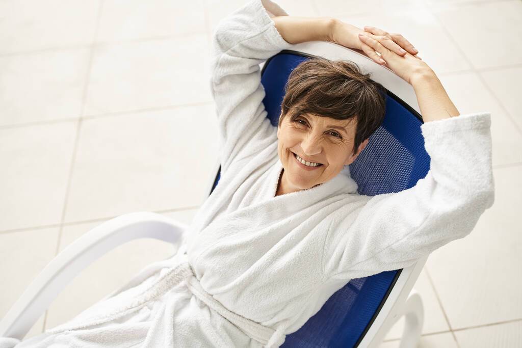 glückliche Frau mittleren Alters mit kurzen Haaren entspannt auf Liege, weißer Robe, Wellness-Center, Blick in die Kamera - Foto, Bild