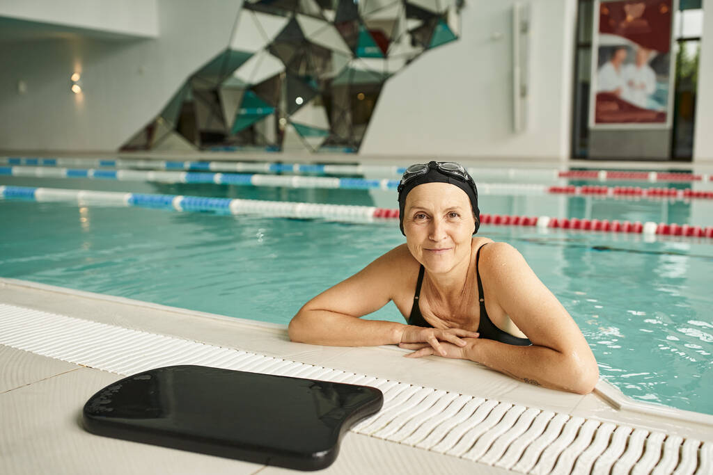 mujer feliz apoyada en la piscina cerca de la tabla flotante, gorra de natación y gafas, sonrisa, mediana edad - Foto, imagen