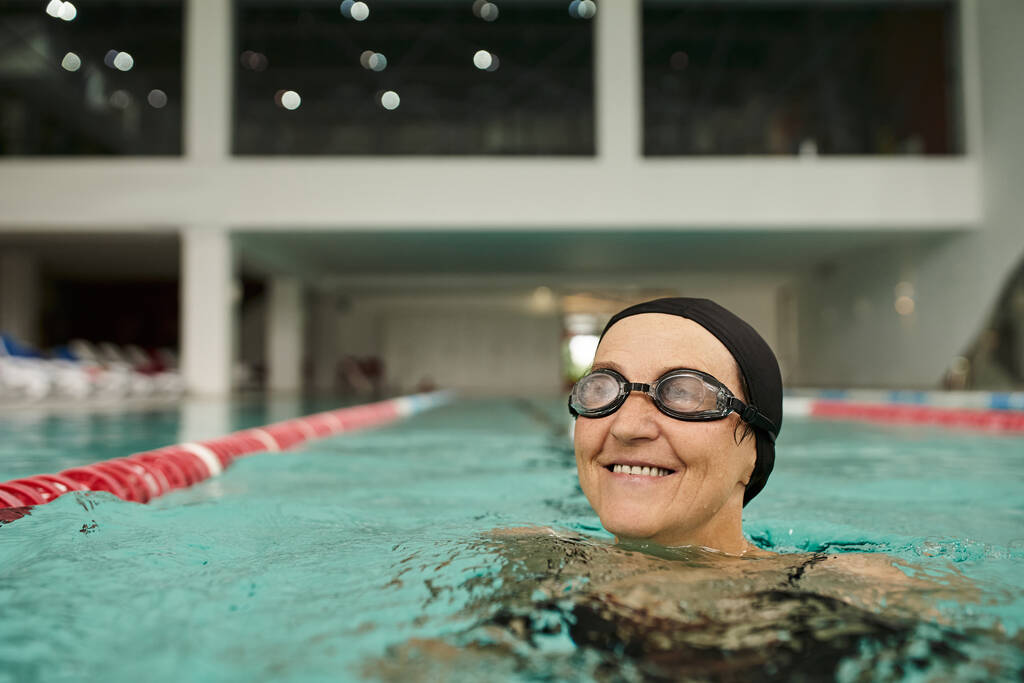 pogodna kobieta w średnim wieku w czapce i okularach kąpielowych w basenie, wodzie, centrum rekreacji, spa - Zdjęcie, obraz