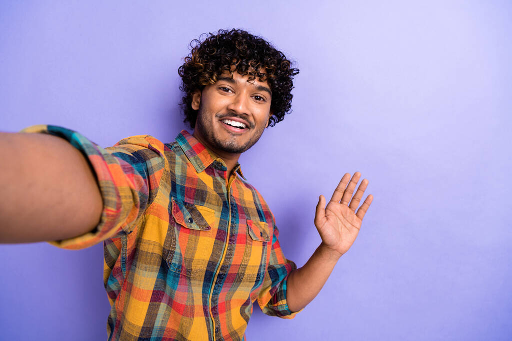 Selfie nuoren miehen arabi kiharat hiukset kuluvat ruudullinen tyylikäs paita heiluttaa kämmen hei ele positiivinen tunnelma eristetty violetti väri tausta. - Valokuva, kuva
