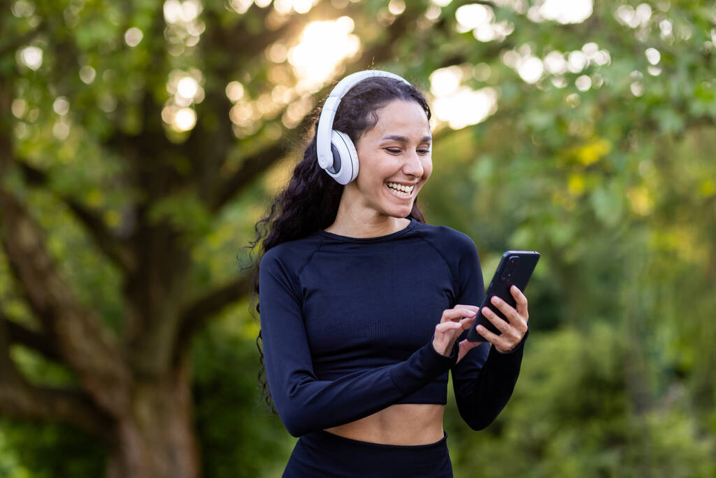 Joven hermosa mujer latinoamericana camina en el parque durante ejercicios físicos activos, mujer utiliza una aplicación para escuchar audiolibros y música en el teléfono, deportista sonriente en auriculares. - Foto, Imagen