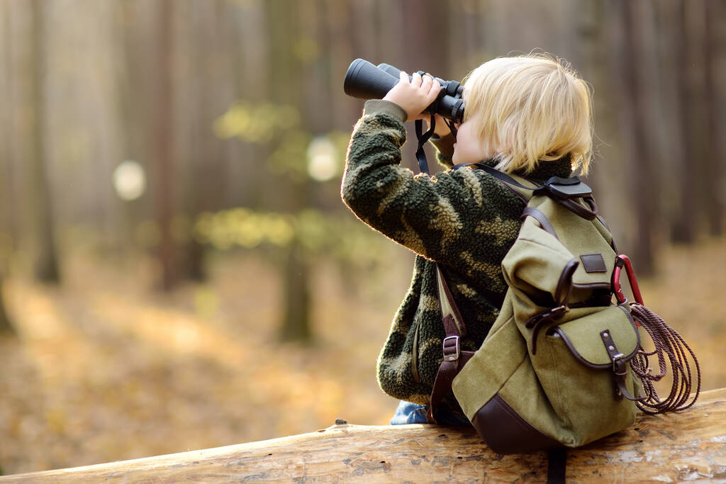 秋の森の中でハイキング中に双眼鏡を持つ小さな男の子のスカウト。子供たちは大きな倒木の上に座って双眼鏡を見ています。子供のための冒険、スカウティング、ハイキング観光の概念. - 写真・画像