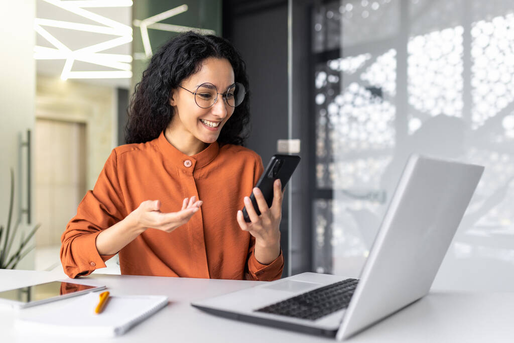 Young beautiful woman talking on a video call using an application on a smartphone, a Hispanic woman is smiling and browsing online internet pages while sitting at a workplace inside an office. - Photo, Image