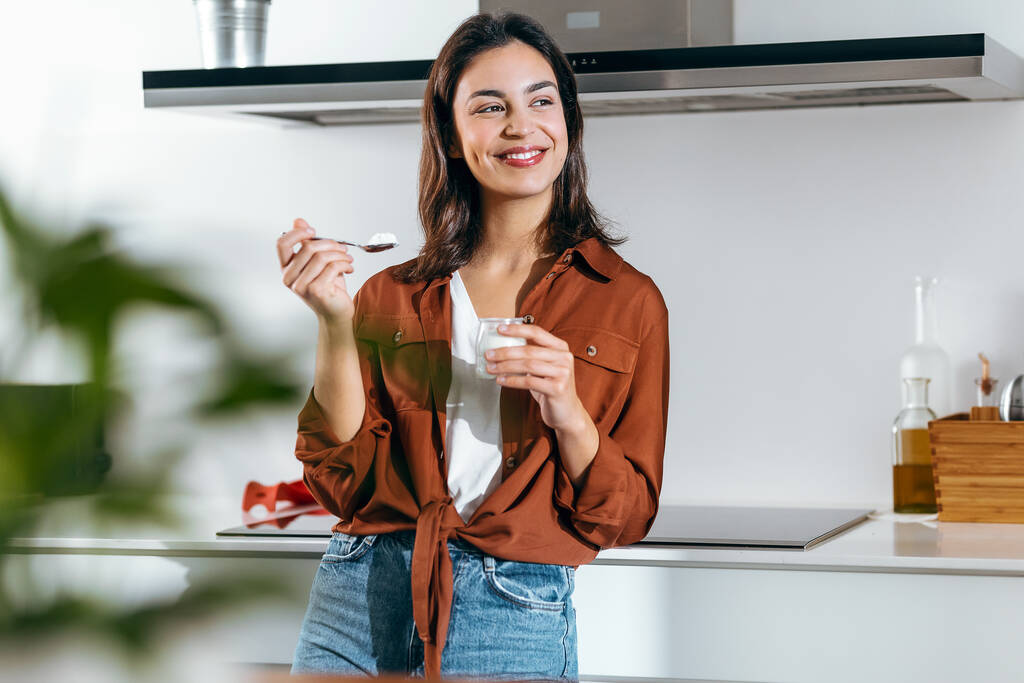 Egy gyönyörű, boldog nő joghurtot eszik, miközben előre néz otthon a konyhában. - Fotó, kép