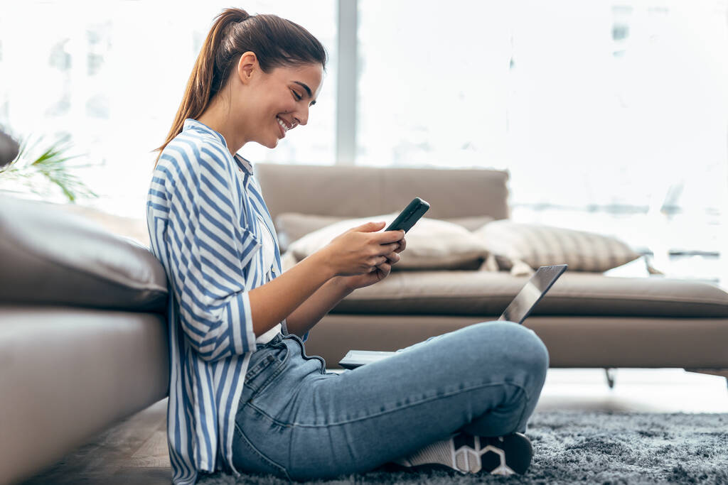 Laukaus kaunis nuori nainen käyttää matkapuhelintaan työskennellessään kannettavan tietokoneen istuu lattialla kotona. - Valokuva, kuva