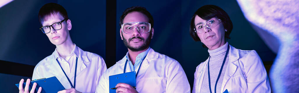 banner, Tres científicos examinan dispositivo recién creado en Neon-Lit Science Center - Foto, Imagen