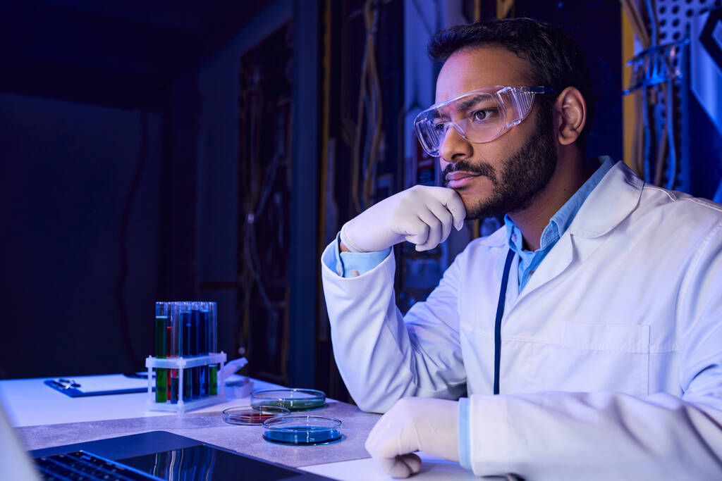 indyjski naukowiec w goglach pracujący na laptopie w pobliżu probówek i płytek Petriego w futurystycznym laboratorium - Zdjęcie, obraz