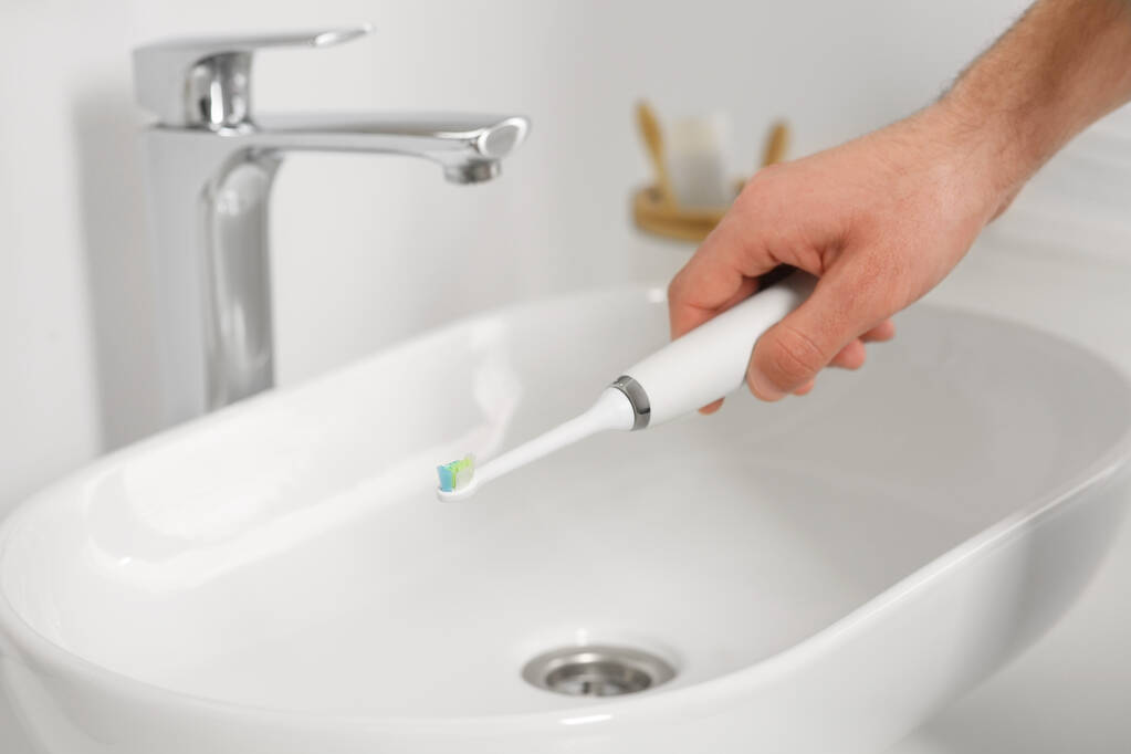 Мужчина держит электрическую зубную щетку над раковиной в ванной, крупным планом - Фото, изображение