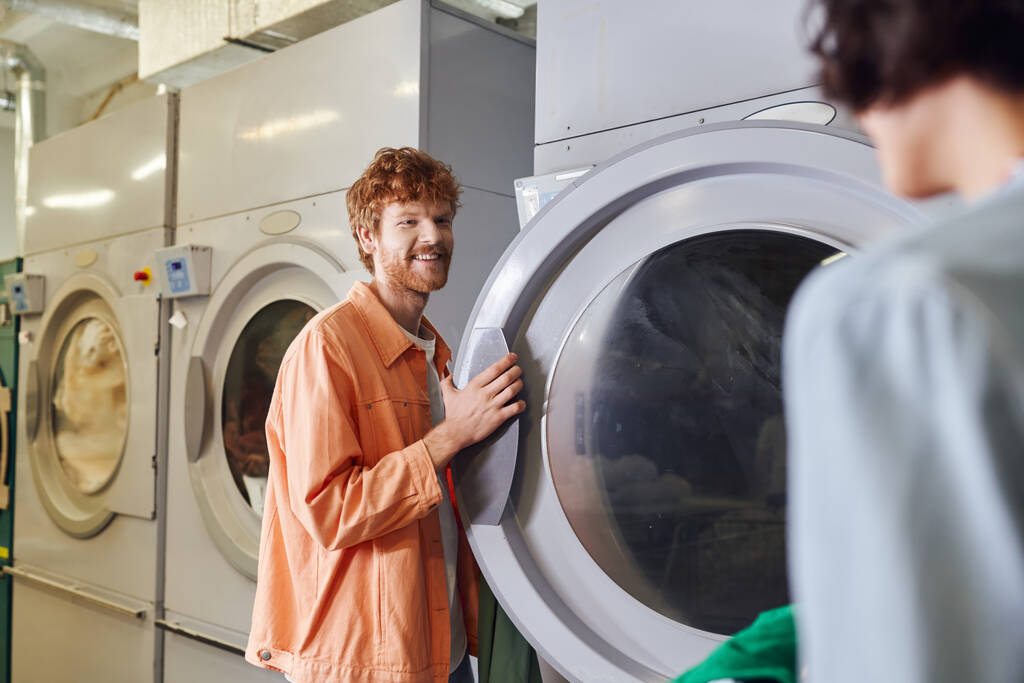 smiling man standing near washing machine and blurred girlfriend with basin in self service laundry - Photo, Image