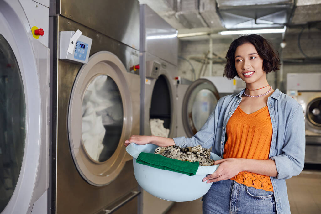 Lächelnde junge Asiatin hält Korb mit Kleidung neben Waschmaschine in Münzwäsche - Foto, Bild