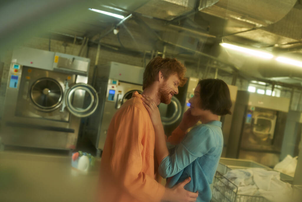 side view of smiling young multiethnic couple touching each other in blurred coin laundry - Photo, Image