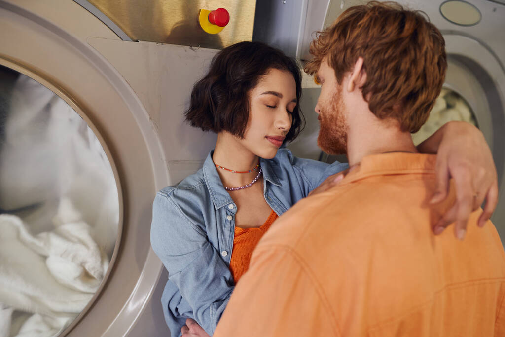 ロマンチックな若いアジアの女性は,公共の洗濯機で洗濯機の近くで赤毛ボーイフレンドを抱擁 - 写真・画像