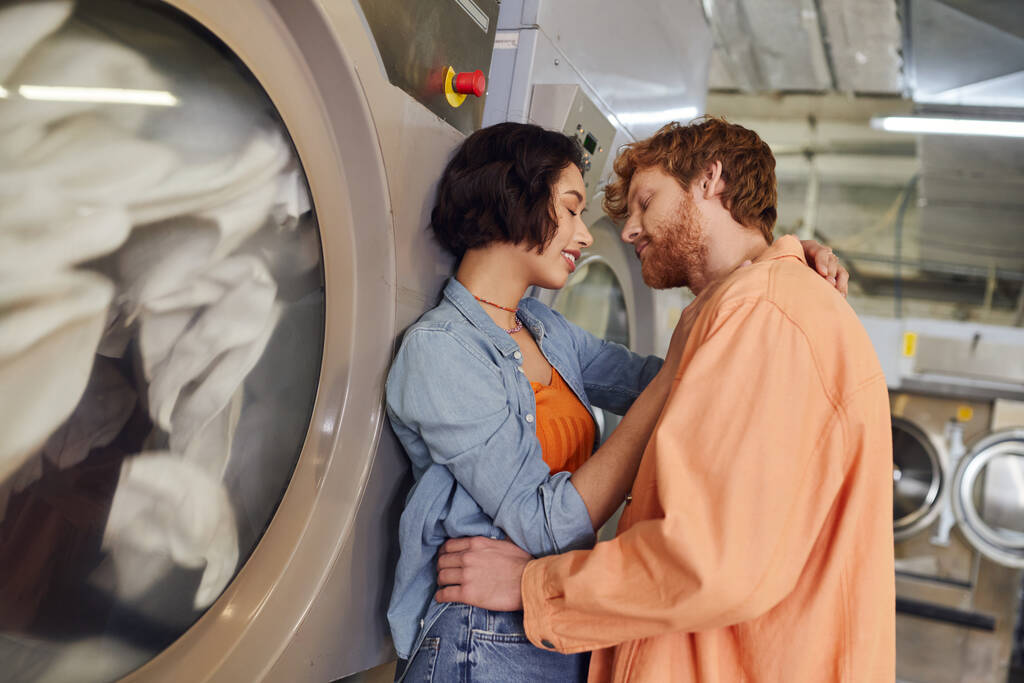 笑顔のアジアの女性は公共の洗濯機の洗濯機の近くで赤毛ボーイフレンドにキス - 写真・画像