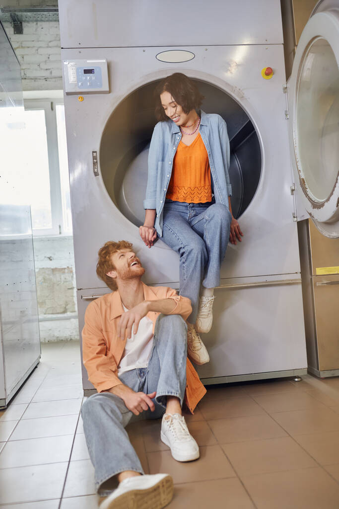 allegro giovane asiatico donna seduta su lavatrice vicino rossa fidanzato in pubblico lavanderia - Foto, immagini