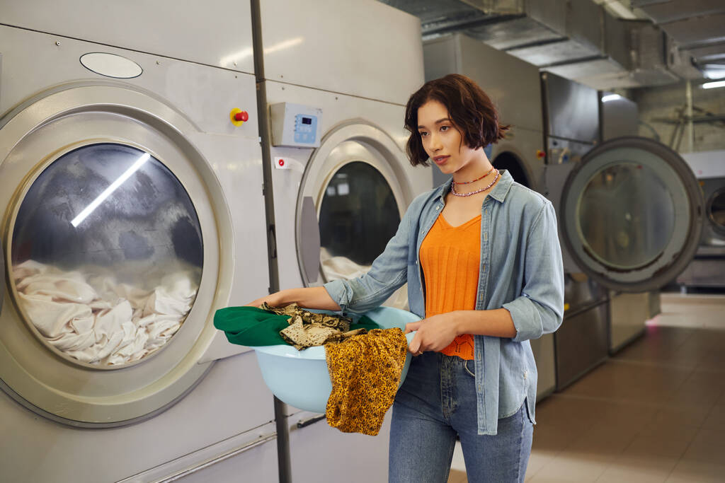 nuori aasialainen nainen tilalla kori vaatteita lähellä pesukoneita julkisessa pyykissä - Valokuva, kuva