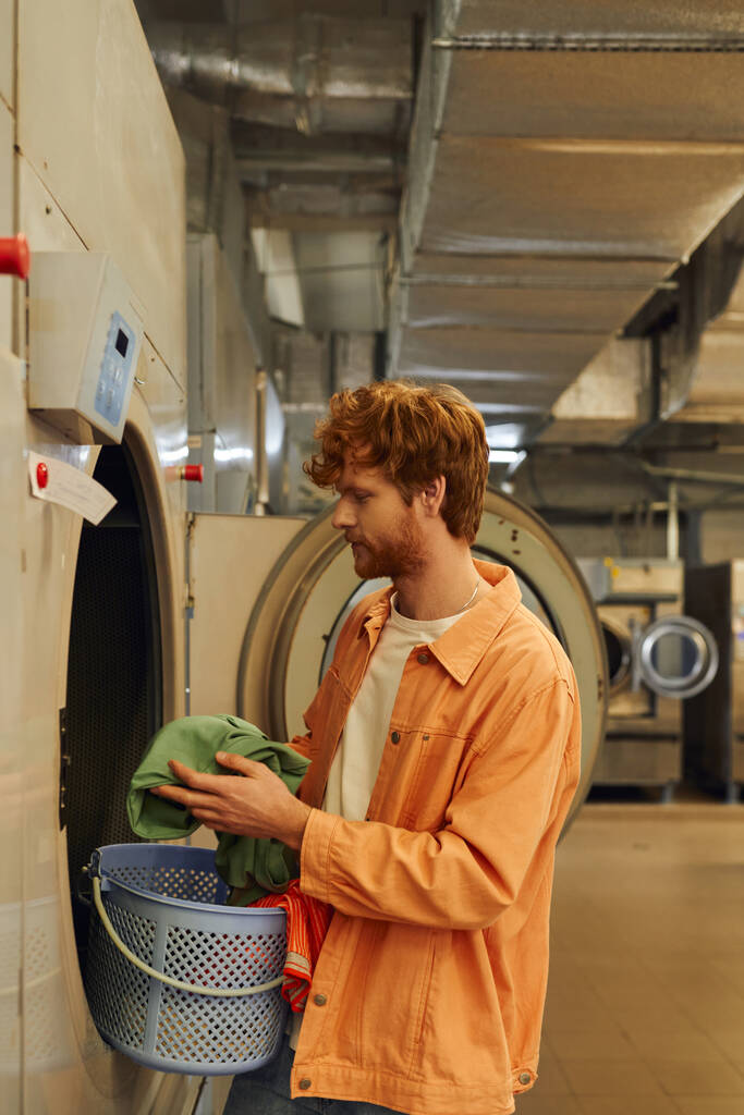 πλαϊνή άποψη του νεαρός κοκκινομάλλης κρατώντας ρούχα κοντά στο πλυντήριο σε δημόσια πλυντήρια - Φωτογραφία, εικόνα
