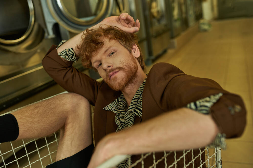 junger stylischer rothaariger Mann in Jacke blickt in die Kamera, während er im Einkaufswagen in der Münzwäsche sitzt - Foto, Bild