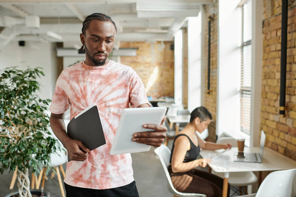 hombre afroamericano usando tableta, carpeta de retención, trabajando cerca de compañera de trabajo, proyecto de startup - Foto, imagen